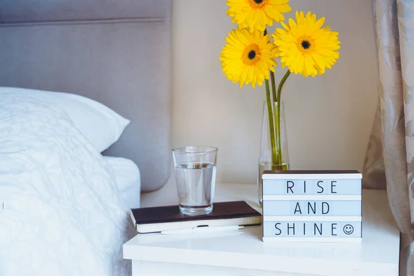 Good morning concept. Inspiration Motivational Life Quotes "Rise and shine" message on a lightbox with notebook and glass of water and fresh sunny yellow gerbera flowers on the bedside table. — Stock Photo, Image