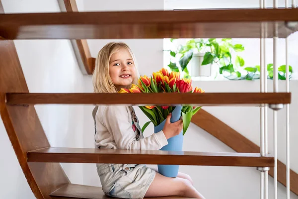 現代の家の木製の階段に座ってチューリップの巨大な束で歯のない少女を笑顔。女の子は花を得る。母の日への贈り物花束誕生日の驚き。幸せな子供時代. — ストック写真