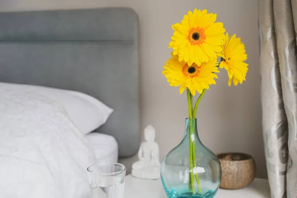 Good morning concept. Sunny yellow gerbera flowers and glass of water on the bedside table. Simple details for good mood for all day. Bedroom Design Ideas. Selective focus, copy space. — Stock Photo, Image