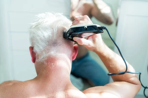 Vista posterior hombre pelo blanco haciendo auto corte de pelo con un clipper y se mira en el espejo. Autocuidado en el hogar en las condiciones de cuarentena global y peluquerías cerradas y salones de belleza. máquina de afeitar eléctrica — Foto de Stock