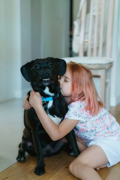 Niña abrazando a una mascota grande perro negro mientras está sentado en el suelo en la sala de estar. Divertirse con animales domésticos. Concepto de animales de amor. Tarjeta vertical. Enfoque selectivo — Foto de Stock