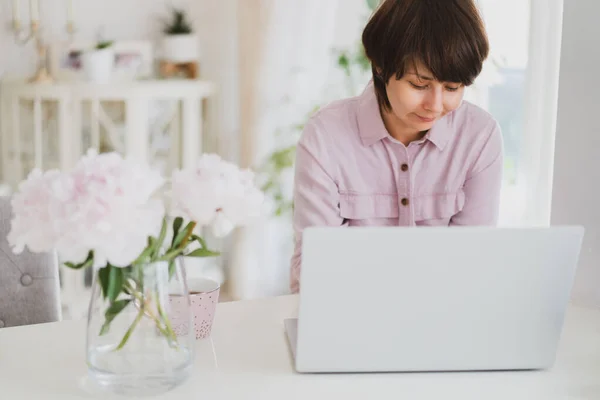 Mulher de raça mista trabalhando em casa usando um laptop. Espaço de trabalho acolhedor com flores de peônias rosa frescas e uma xícara de chá na mesa branca com fundo interior claro. Trabalho remoto em casa. Espaço de cópia — Fotografia de Stock