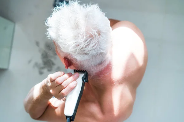 Vista superior hombre pelo blanco haciendo auto corte de pelo con un cortador y se mira en el espejo. Autocuidado en el hogar en las condiciones de cuarentena global y peluquerías cerradas y salones de belleza. máquina de afeitar eléctrica — Foto de Stock