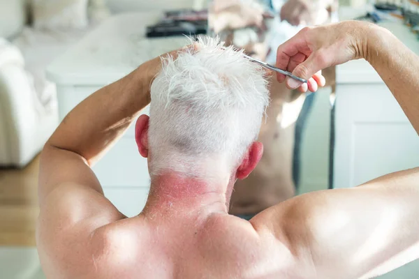 Πίσω όψη λευκό μαλλιά άνθρωπος κάνει αυτο-κούρεμα με ψαλίδι και κοιτάζει στον καθρέφτη. Αυτοφροντίδα στο σπίτι στις συνθήκες της παγκόσμιας καραντίνας και κλειστά κομμωτήρια και ινστιτούτα αισθητικής. — Φωτογραφία Αρχείου