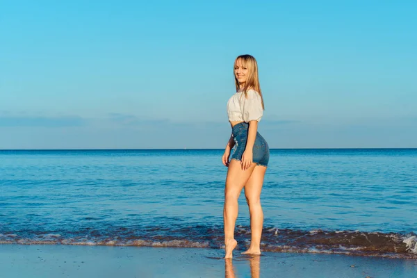 하얀 위와 청바지 반바지를 입은 건강 한 여성 이 해변에서 산책을 하는 동안푸른 바 다 와 하늘 배경 위에서 웃고 있다. 여름 방학, 휴가. 복사 공간. — 스톡 사진