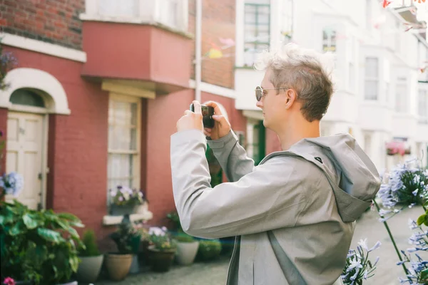 Jovem turista filmando vídeo ou tirar fotos no celular durante a caminhada na área residencial cottagecore. Bela vista de rua com muitas plantas e flores em vasos. — Fotografia de Stock