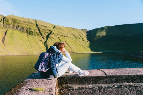 뒤를 돌아보면, 산 호숫가 근처에 배낭을 메고 앉아 푸른 언덕 - 등산 목적지 - 의경 치를 즐기고 있는 여자가 보인다. 여행하는 생활 방식은 모험을 선호 합니다. 자연 에서의 휴가. 자유와 평온 — 스톡 사진