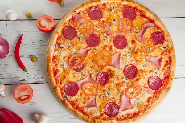 Close up Delicious Meat Pizza with Salami, Bacon and Tomato Slices on a cutting board on white wooden background with different colourful vegetables top view