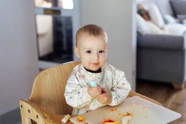 Cute baby learning to eat food with baby spoon by himself on high chair baby led weaning or blw. Infant self-feeding solid food, making mess, fine motor development. Kid enjoying healthy nutrition
