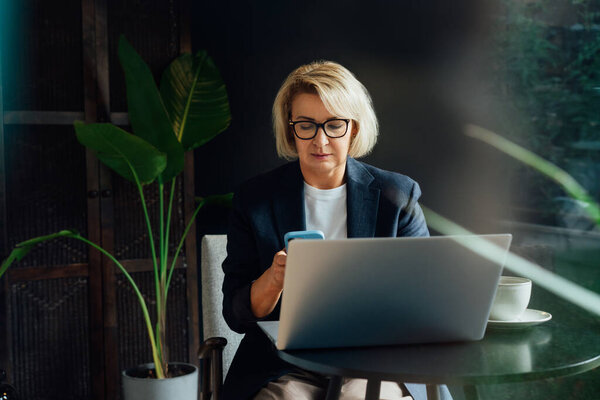 Stylish 50s confident mature businesswoman, middle aged company ceo director, experienced senior female professional, business coach using phone and working on laptop in office. Female leader.