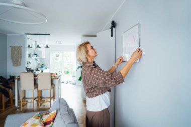 Side view middle aged woman putting painting picture in frame on the wall. Casual lady taking care of coziness in her new stylish apartment. Mature female decorating room. Moving and renovate house.
