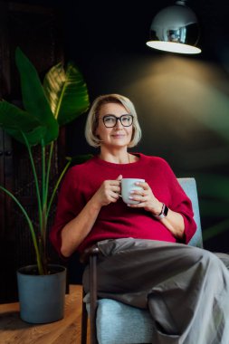 Orta yaşlı bir kadın Hygge tarzı iç mekanında bir fincan sıcak içecekle rahatlıyor. Rüya gören bayan, stres olmadan sakin havanın tadını çıkar, yalnız olmanın tadını çıkar. Biraz ara ver ve rahatla. Sıcak sonbahar ya da kış hafta sonu