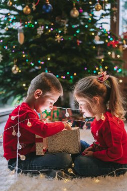 Evde Noel ağacının altında oturan iki neşeli süveter giymiş gülen çocuk. Duygusal kız ve erkek kardeş Noel hediyelerini açmak için heyecanlı hissediyorlar, eğleniyorlar, çocukluk mucizesi konsepti.