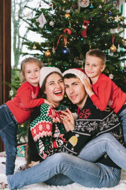 Noel 'de Noel ağacının altında hediyelerle oturmuş neşeli süveterlerle gülümseyen aile fotoğrafı. Mutlu ebeveynler ve çocuklar sarılıyor, heyecanlı hissediyorlar. Ailecek vakit geçireceğiz. Mutlu çocukluk.