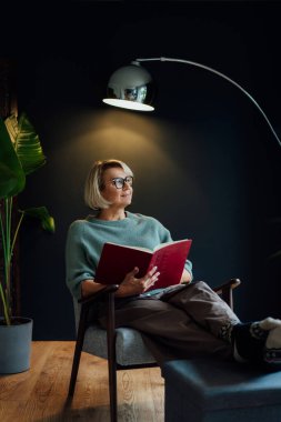 Orta yaşlı bir kadın kitap okuyor. Skandal tarzda. Karanlık bir iç mekan. Rüya gören bayan, stres olmadan sakin havanın tadını çıkar, yalnız olmanın tadını çıkar. Biraz ara ver ve rahatla. Evde rahat bir tatil zamanı. Dikey.