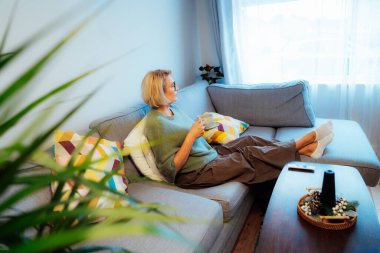 Orta yaşlı bir kadın Hygge Home 'daki kanepede bir fincan sıcak içecekle rahatlıyor. Rahat kış ya da sonbahar hafta sonları. Biraz ara ver ve rahatla. Hanımefendi sakin ve yalnız olmanın keyfini çıkarın..