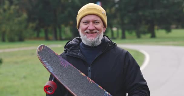 Retrato de un hombre de pelo gris adulto en un parque de la ciudad, un hombre sosteniendo un longboard en sus manos y sonriendo, una pista de esquí sobre ruedas en un parque de la ciudad. — Vídeos de Stock