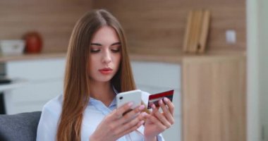 Genç bir kadın oturma odasındaki kanepede oturuyor ve internetten akıllı telefon kullanarak satın alımları ödüyor..