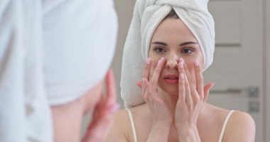 Genç bayan banyoda aynanın önünde duruyor ve yüzüne krem sürüyor, cilt bakımı..