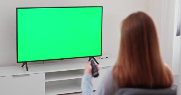 Girl is sitting on the sofa in the living room and watching TV, green screen, the girl switches channels using the remote control, chroma key template. — Stock Video