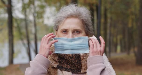 Portret van een volwassen vrouw in een bos Park, oudere vrouw zet op beschermende medische masker, herfst stemming, grijs-harige vrouw in een stad Park, wazig achtergrond. — Stockvideo