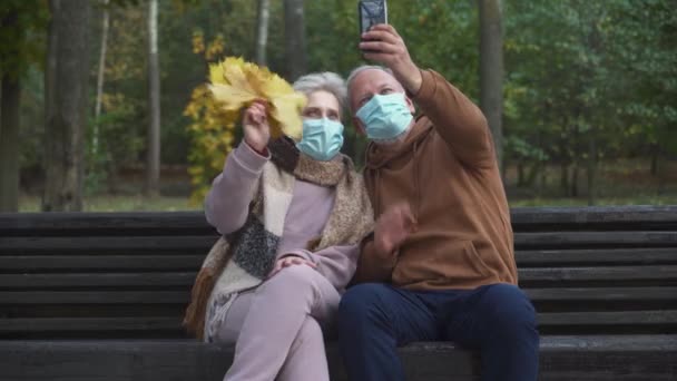 Ouderen in medische maskers zitten op een bankje in een bos Park en nemen een selfie video op een smartphone, grijs haar man en een vrouw communiceren via conference call, leven in een pandemie. — Stockvideo