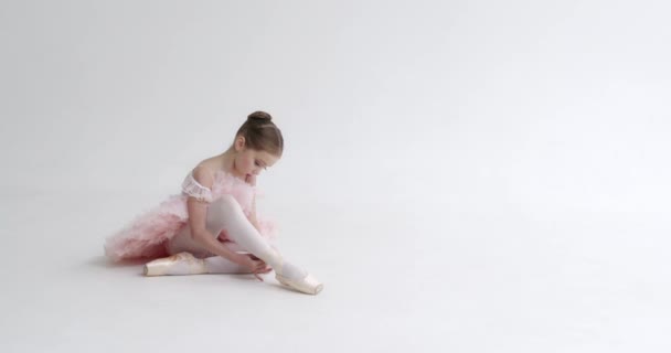 Mała dziewczynka w białej spódniczce siedzi na podłodze i wiąże wstążki w butach Pointe, młoda balerina przygotowująca się do treningu, białe tło. — Wideo stockowe