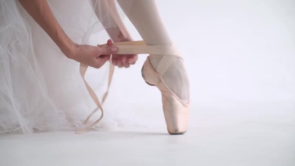 하얀 턱시도를 입은 암컷이 바닥에 앉아 그녀의 푸네 테 신발의 리본을 매고 있습니다. 젊은 발레리나는 하얀 배경에서 공연을 준비하고 있습니다.. — 비디오