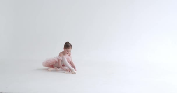 Niña en un tutú blanco se sienta en el suelo y ata las cintas de sus zapatos Pointe, joven bailarina, fondo blanco. — Vídeos de Stock