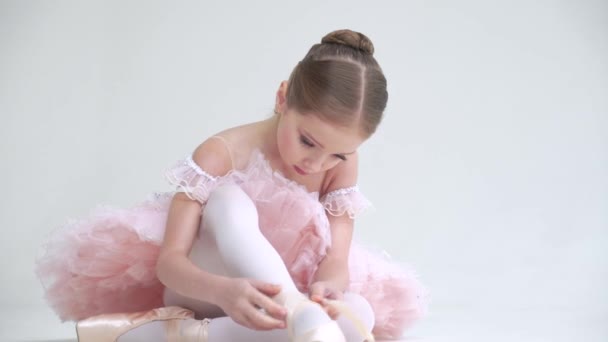 Niña en un tutú blanco se sienta en el suelo y ata las cintas de sus zapatos Pointe, joven bailarina sobre un fondo blanco. — Vídeo de stock