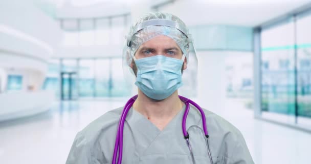 Portré egy férfi orvos védőfelszerelés és orvosi maszk, férfiak műanyag napellenző belenéz a kamerába, védelem egy covid-19, az orvos áll a háttérben egy modern kórház, elmosódott — Stock videók