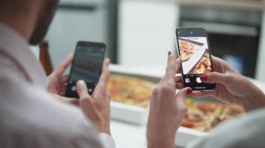 Ev partisi, mutfakta masada oturan ve akıllı telefonlardan pizza fotoğrafı çeken genç bir çift..