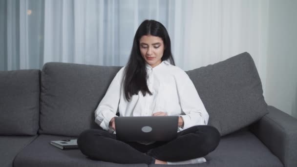 Jonge vrouw freelancer zit op de bank en werkt aan een laptop, typt tekst op het toetsenbord, 's avonds tijd, werk op afstand van huis. — Stockvideo