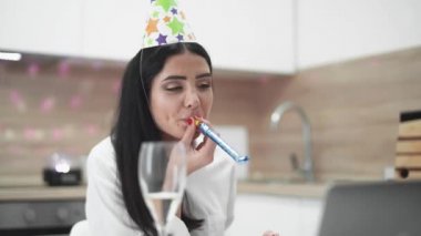 Neşeli bir ruh hali, dizüstü bilgisayarda mutfakta oturan ve doğum gününü online kutlayan bir kız, tatil için arkadaşlarıyla görüntülü konuşma, bir kız şarap içiyor ve laptop kamerasına bir hediye gösteriyor, olumlu duygular.