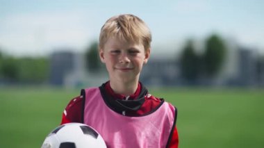 Futbolcu bir çocuğun portresi, genç bir çocuk futbol sahasının yanında duruyor ve kameraya bakıyor, elinde bir futbol topu tutuyor, geçmişi bulanık..