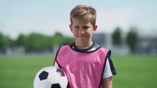 Portrét chlapce fotbalisty, mladého chlapce stojícího u fotbalového hřiště a dívajícího se do kamery, držícího v rukou míč, rozmazané pozadí. — Stock video