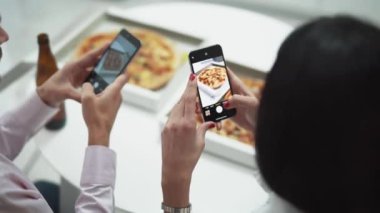 Ev partisi, mutfakta masada oturan ve akıllı telefonlardan pizza fotoğrafı çeken genç bir çift..