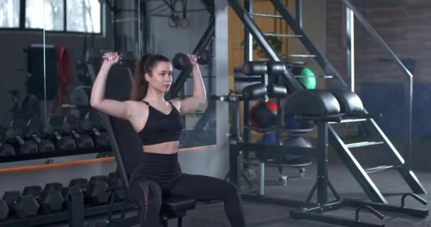 Fiatal nő edzés a tornateremben, a lány súlyzókat emel fel, miközben a padon ül, izomedzések súlyokkal, 4k 50fps. — Stock videók