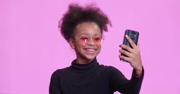 Retrato de una hermosa chica africana sobre un fondo rosa, la chica toma video de sí misma en un teléfono inteligente, video blogger hablando en una cámara, emociones positivas, 4k 50fps. — Vídeo de stock