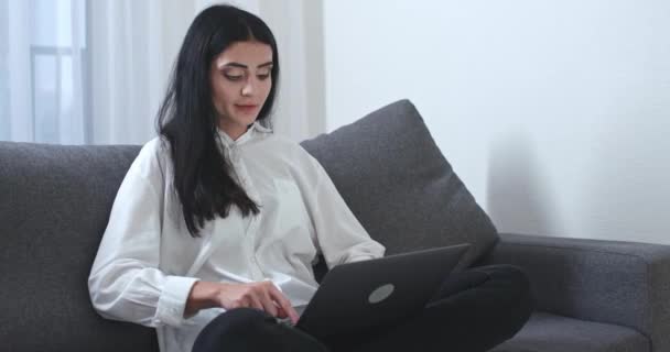 Serbest çalışan bir kadın kanepede oturuyor ve dizüstü bilgisayarda çalışıyor, klavyeye mesaj yazıyor, akşam vakti, evden uzaktan kumandayla.. — Stok video