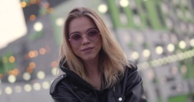 Genç, güzel bir kız gülümsüyor ve kamerada poz veriyor, modern bir yerleşim yerinin yakınında, şehirde bir akşam, arka planda bulanık ışıklar, pozitif duygular..