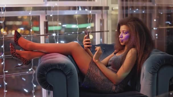 Glamouröse Afrikanerin liegt auf einem Stuhl im Loungebereich und benutzt Smartphone, Online-Kommunikation, Mädchen langweilt sich auf einer Party. — Stockvideo