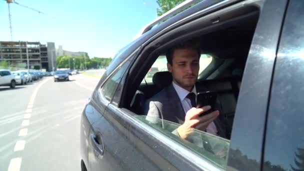 Joven hombre de negocios está sentado en un coche en movimiento y hablando en un teléfono móvil, hombre gerente superior en un traje está sentado en la parte posterior de un coche. — Vídeos de Stock