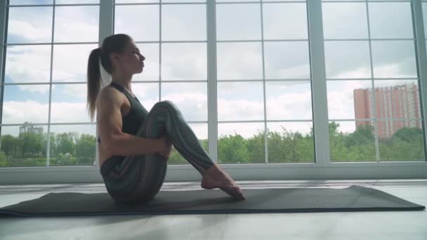 Ung kvinna som gör yoga i ett vitt rum fyllt med ljus, flickan utför yoga står nära det stora fönstret. — Stockvideo