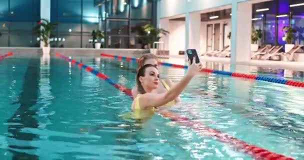 Young females in the pool, two beautiful females in yellow swimsuits take a selfie on a smartphone while in the pool. — Stock Video