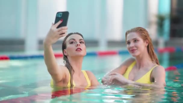 Jonge vrouwen in het zwembad, twee mooie meisjes in gele zwempakken nemen een selfie op een smartphone. — Stockvideo