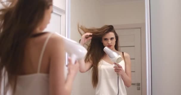 Joven bonita hembra se para cerca del espejo y se seca el cabello con un secador de pelo, vista del reflejo en el espejo. — Vídeos de Stock