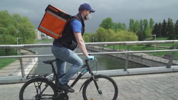 Entrega de alimentos nas condições de uma pandemia viral, correio em máscara médica com uma mochila térmica monta uma bicicleta para o cliente. — Vídeo de Stock