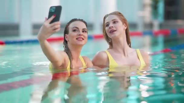 Молодые женщины в бассейне, две красивые девушки в жёлтых купальниках делают селфи на смартфоне, девушки улыбаются и позируют перед камерой. — стоковое видео