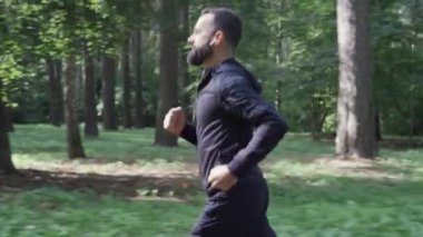 Yetişkin bir adam yaz ormanlarında koşuyor, kamera takip ediyor, kablosuz kulaklık takıyor, sağlıklı bir yaşam tarzı var..